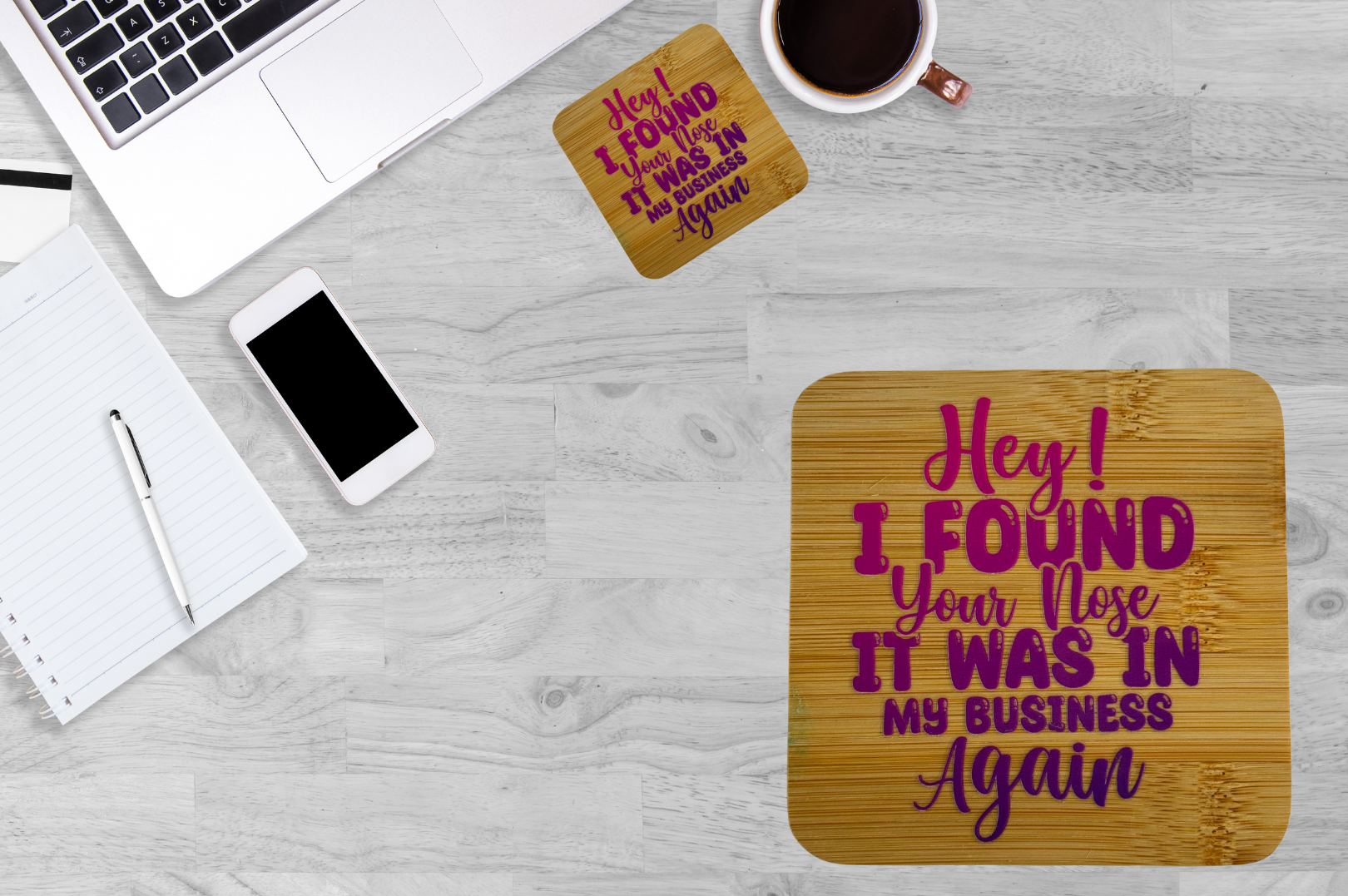 A flat-lay view of a light gray wood-grain desk featuring two square coasters with a humorous quote in pink and purple lettering. The main coaster in the foreground reads 'Hey! I FOUND Your Nose.