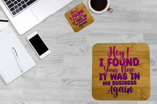 A flat-lay view of a light gray wood-grain desk featuring two square coasters with a humorous quote in pink and purple lettering. The main coaster in the foreground reads 'Hey! I FOUND Your Nose.